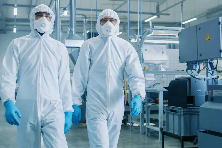 Two people wearing full-body white protective suits, gloves, and face masks walk through an industrial laboratory or manufacturing facility.