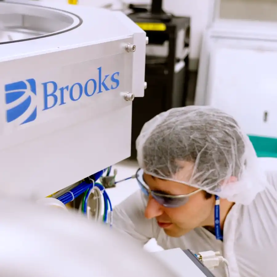 A person wearing protective clothing and goggles inspects equipment labeled "Brooks" in a laboratory or cleanroom environment.