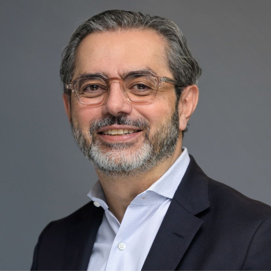 A middle-aged man with glasses, gray hair, and a beard, wearing a dark suit jacket over a light-colored shirt, poses against a plain gray background.