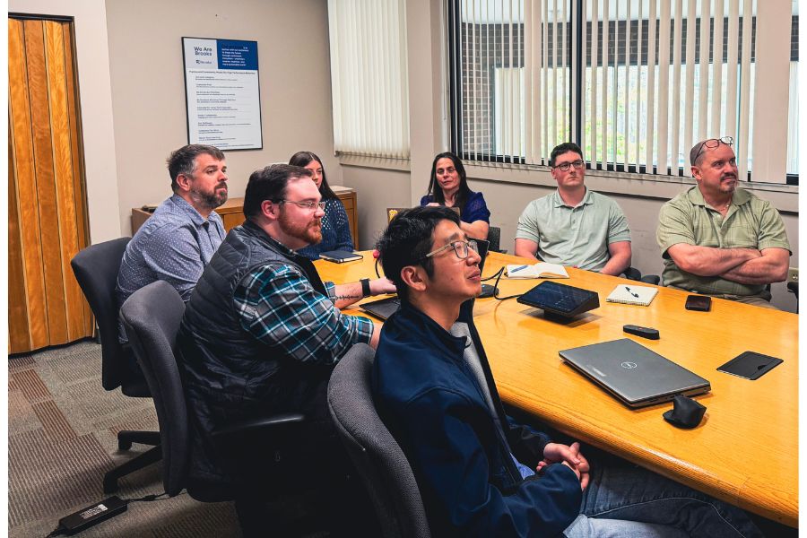 Seven people sit around a conference table in an office, attentively watching something off camera; laptops, notebooks, and papers are on the table.