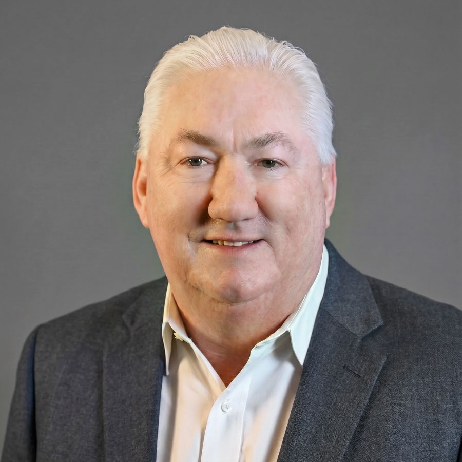 A middle-aged man with short white hair wearing a dark blazer and white collared shirt poses in front of a plain gray background.