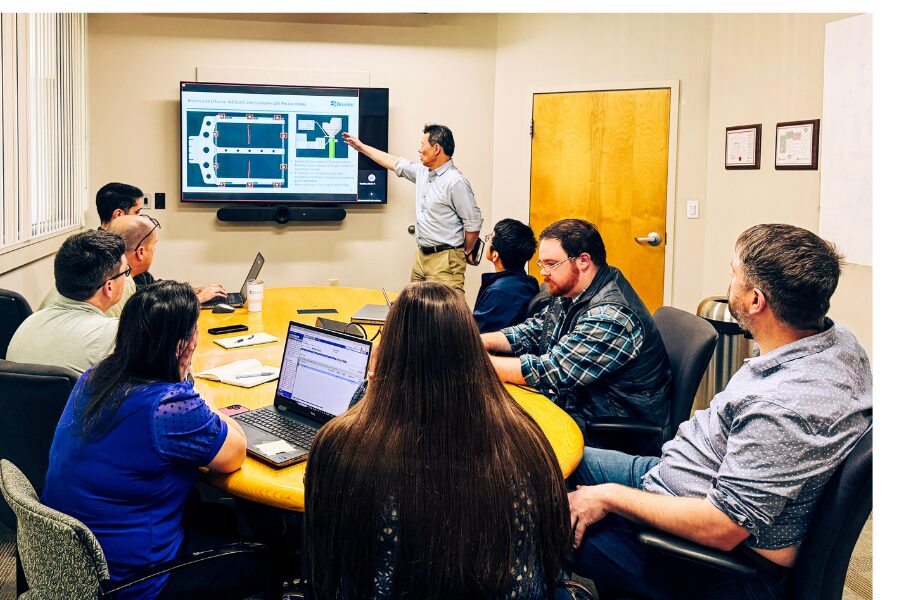 A man stands and points at a presentation on a screen while six people seated around a conference table listen and take notes on laptops.